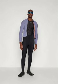 Férfi sportoló fekete, teljes testet takaró kerékpáros ruhát visel, világoslila kerékpáros dzseki narancssárga cipzárral, fekete cipőket és nagy kék sport napszemüveget.