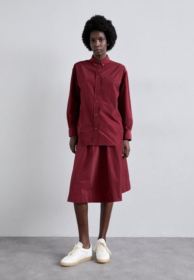 Oversized burgundy shirt, button-up with pocket, paired with a matching knee-length skirt. White sneakers with beige soles. Smooth cotton texture.