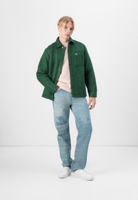 Veste en mélange de laine verte avec un devant à boutons, une poche poitrine unique, portée sur un pull rose pâle, associée à un jean déchiré bleu clair et des baskets blanches.