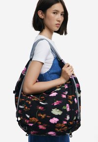 Black floral shoulder bag with pink, white, and green flower patterns. Features striped handles and a textured surface. Adjustable straps included.