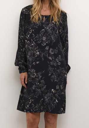 Žena v volnějším černém šatu s dlouhými rukávy a šedými květinovými vzory, stojící před jednoduchým bílým pozadím.