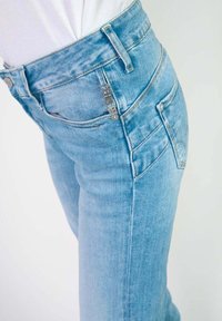 Jean en denim bleu clair avec une coupe slim, doté d'une taille haute, de coutures détaillées et d'un logo discret sur la poche avant.
