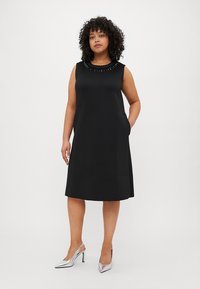 Μαύρο αμάνικο φόρεμα με στρογγυλή λαιμόκοψη διακοσμημένο με λεπτομέρειες από χάντρες, με πλαϊνές τσέπες και χαλαρή σιλουέτα Α. Ασημένια ψηλοτάκουνα παπούτσια.