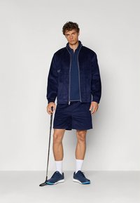 Navy corduroy bomber jacket, matching shorts, and blue athletic shoes. Model holds a golf club. Simple design and textured fabric.