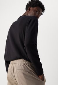 Un pull noir avec des poignets côtelés associé à un pantalon beige à taille élastique, avec des poches latérales et une texture lisse. Design minimaliste.