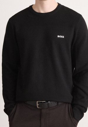 Osoba ubrana w czarną koszulę z długim rękawem z logo "BOSS" na piersi, ciemne spodnie i czarny pasek, z rękami w kieszeniach.