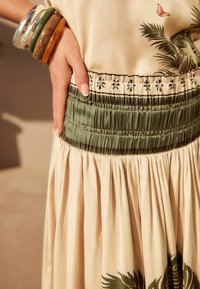 Falda beige claro con cintura elástica verde, con bordado floral. Textura suave, pliegues sueltos y detalle de diseño botánico.