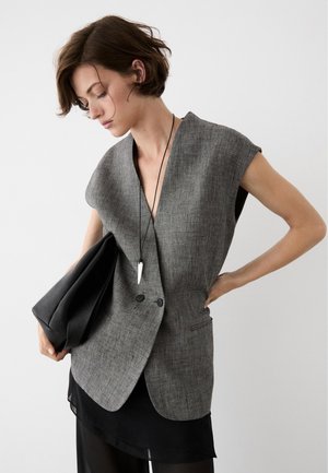 Kobieta o krótkich brązowych włosach, ubrana w szarą kamizelkę bez rękawów, czarną warstwową spódnicę, długi naszyjnik z zawieszką, trzymająca dużą czarną kopertówkę.