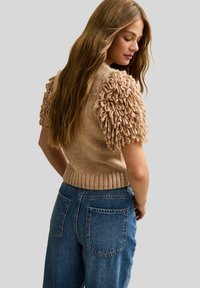 Knit sweater in beige with textured, fluffy short sleeves. Paired with high-waisted blue denim jeans. Rear view against a light background.