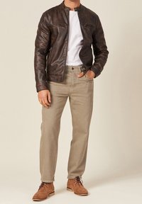 Man met een bruine leren jas, wit T-shirt, beige broek en bruine suède veterschoenen, staand met één hand in de zak.