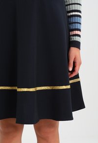 Robe noire avec une jupe évasée, dotée d'une bande pailletée dorée et d'accents de manches rayées multicolores. Tissu lisse, design à longueur de genoux.