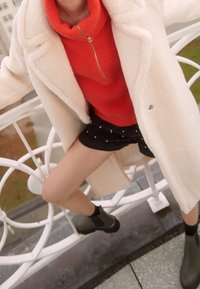 Abrigo grande de piel sintética blanca, suéter rojo con cremallera en canalé, pantalones cortos negros con detalles de perlas y botines verde oscuro.