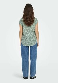 Blusa verde a maniche corte con motivo floreale bianco, orlo arrotondato, abbinata a jeans azzurri a vita alta con gamba dritta e scarpe nere.