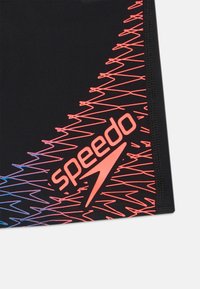 Melnas peldbikses ar rozā zigzaga rakstu un "speedo" logo. Materiāls ir gluds un elastīgs; dizains ir sportisks un piegulošs.