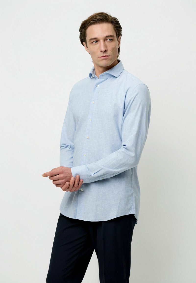 Camisa de vestir ajustada de color azul claro en una tela suave, con cuello abotonado, mangas largas y dobladillo curvado, combinada con pantalones oscuros.