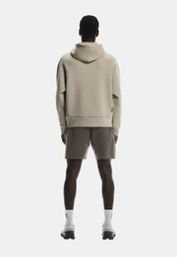 Sweat à capuche beige avec une coupe décontractée, associé à un short marron. Le modèle porte des chaussettes blanches et des chaussures de course noires, vu de derrière.
