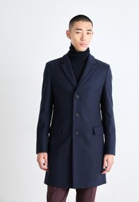 Marineblå ullfrakk med skreddersydd design, enkeltspent, fem knapper og to frontlommer, brukt over en svart høyhalset genser.