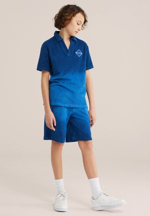 Enfant aux cheveux bouclés portant un polo bleu et un short assorti, regardant vers le bas, debout devant un fond beige uni.