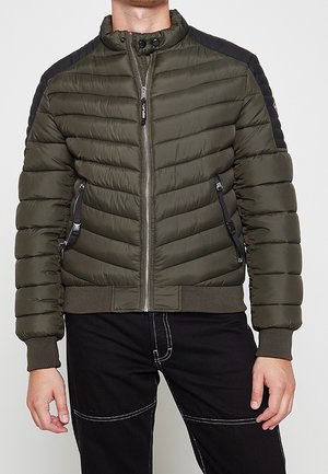 Mies, joka pukeutunut oliivinvihreään tikattuun toppatakkiin, jossa on mustat olkatyynyt, edessä vetoketju, sivuilla vetoketjulliset taskut sekä resorit hihansuissa ja helmassa.