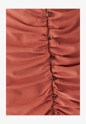 Punakaspruun kogutud kangas vertikaalsete voldikute ja tumeda pruuni nööbiga kinnitustega. Sile tekstuur, millel on peened mustrid.