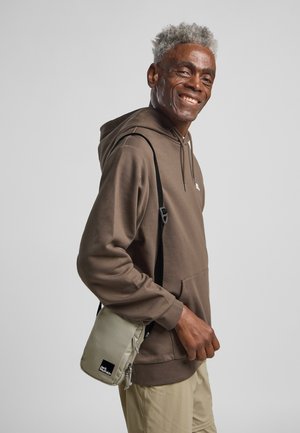 Felpa grigia con cappuccio abbinata a una borsa a tracolla color beige chiaro. La borsa presenta una cerniera e un'etichetta con logo. Vestibilità casual e rilassata.