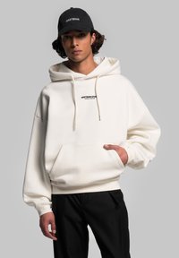 Felpa bianca oversize con tasca frontale e cappuccio con cordino, caratterizzata da un piccolo logo nero sul petto; abbinata a pantaloni neri.