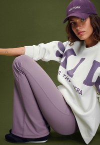Bela športna majica s vijoličnim napisom, v kombinaciji z lilas širokimi hlačami. Model nosi vijolično kapo in črne čevlje, sedi ob zeleni ozadju.
