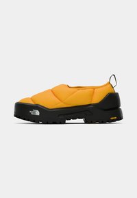Chaussures slip-on matelassées jaunes avec une semelle Vibram noire, présentant une partie supérieure texturée et un onglet au talon noir. Le logo de la marque est affiché sur le côté.