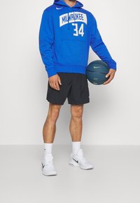 Modrá mikina s bílým potiskem "Milwaukee 34", černé šortky a bílé boty Nike. Drží modrý basketbal a stojí na jednolitém pozadí.