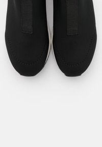 Svarta sneakers med en slät textilövre, med elastiska sidor och rundad tådesign, på en vit sula.