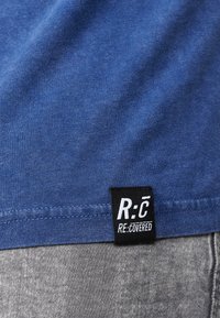 Blå bomullströja med en mjuk textur. Har en svart etikett med "R:C RE.COVERED" vid nederkanten, sydd med en snygg finish.
