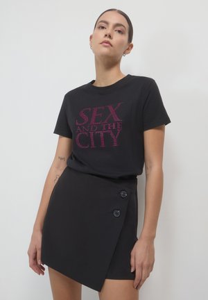 Frau, die ein schwarzes "Sex and the City"-T-Shirt und einen asymmetrischen, geknöpften schwarzen Minirock trägt, vor einem schlichten weißen Hintergrund steht.