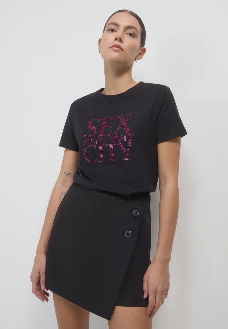 Frau, die ein schwarzes "Sex and the City"-T-Shirt und einen asymmetrischen, geknöpften schwarzen Minirock trägt, vor einem schlichten weißen Hintergrund steht.