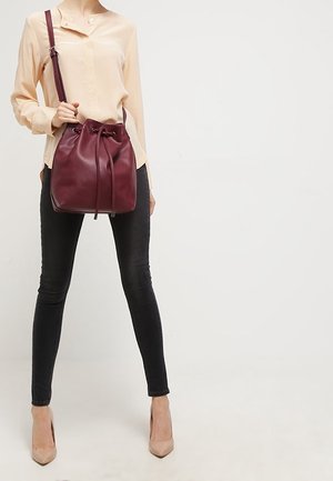 Kobieta ubrana w beżową bluzkę, czarne obcisłe dżinsy i cieliste szpilki, trzymająca bordową skórzaną torbę typu bucket z sznurkiem i paskiem na ramię.