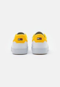 White sneakers with yellow accents, suede and leather materials, rounded shape, textured soles, and a small logo on the heel.