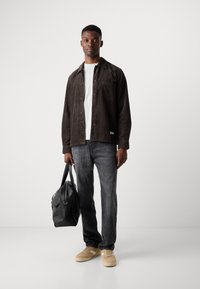 Black corduroy shirt, white T-shirt, gray jeans, tan sneakers, holding a black duffel bag. Simple design, no prominent patterns or accents.