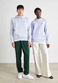 Fatos de treino lilás claros com bolsos frontais e cordões, combinados com calças verdes e ténis brancos. A mulher usa calças largas em tom creme.