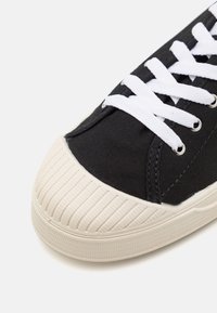 Svart kanvas sneakers med vit gummitå och snören. Har texturerade sidopaneler och en randig gummisul.