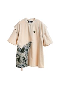 Camisa de manga corta beige con cuello redondo, logo circular negro en el pecho y un parche irregular de color azul-gris verdoso en la parte inferior izquierda.