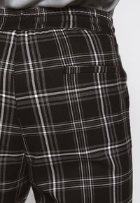 Zwart-wit geruite broek met elastische tailleband, gedragen met een ingestopte grijze shirt, waarin de details van de achterzak zichtbaar zijn.