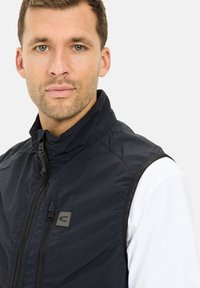 Schwarze Weste mit hohem Kragen und Reißverschluss vorne. Verfügt über ein kleines Logopatch auf der Brust und ein kontrastierendes weißes Hemd darunter.