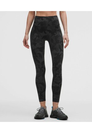 Femme portant des leggings camouflage noirs taille haute et des chaussures de course grises, debout devant un fond clair uni.