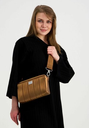 Mustaan mekkoon pukeutunut nainen pitää pronssinväristä olkalaukkua, jossa on leveä remmi, yksinkertaisen valkoisen taustan edessä.