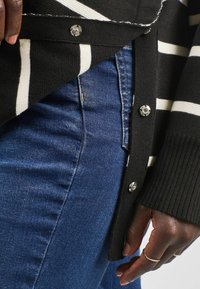 Svartvit randig kofta med silvriga knappdetaljer, parat med mörkblå denim, som visar en närbild av tygstrukturer.