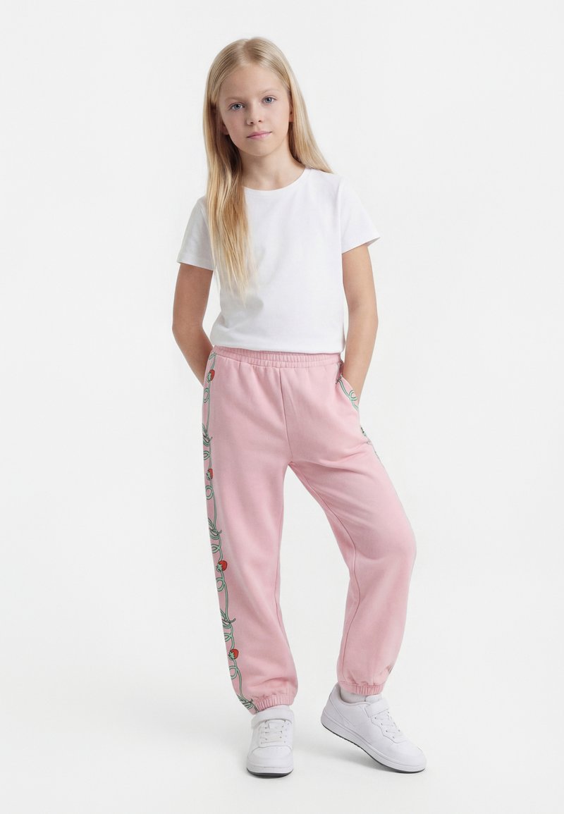 Fiatal lány áll, kezét a zsebében tartja, fehér pólót, rózsaszín melegítőnadrágot virágmintás oldalsó csíkokkal és fehér tornacipőt visel, egyszínű háttér előtt.