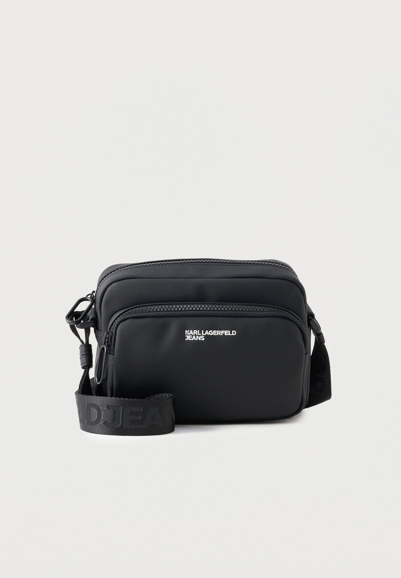 Μαύρη ορθογώνια τσάντα crossbody με δύο θήκες με φερμουάρ, λογότυπο "KARL LAGERFELD JEANS" και ρυθμιζόμενο ιμάντα με λογότυπο, σε λευκό φόντο.