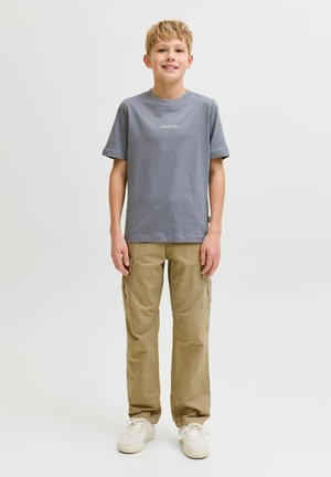 Garçon blond debout, portant un t-shirt gris à manches courtes avec texte, pantalon cargo kaki et baskets blanches sur un fond blanc uni.