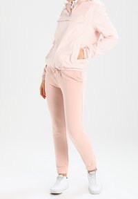 Světlá růžová větrovka se zipem na krku a kapsou vpředu, doplněná o růžové tepláky a bílé tenisky. Jemná textura.