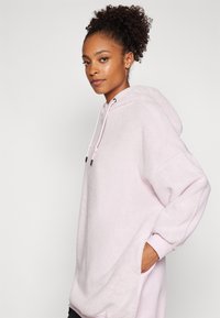 Lys rosa oversized hettegenser laget av myk, teksturert stoff, med snor i hetten, kengurulomme og vide ermer.