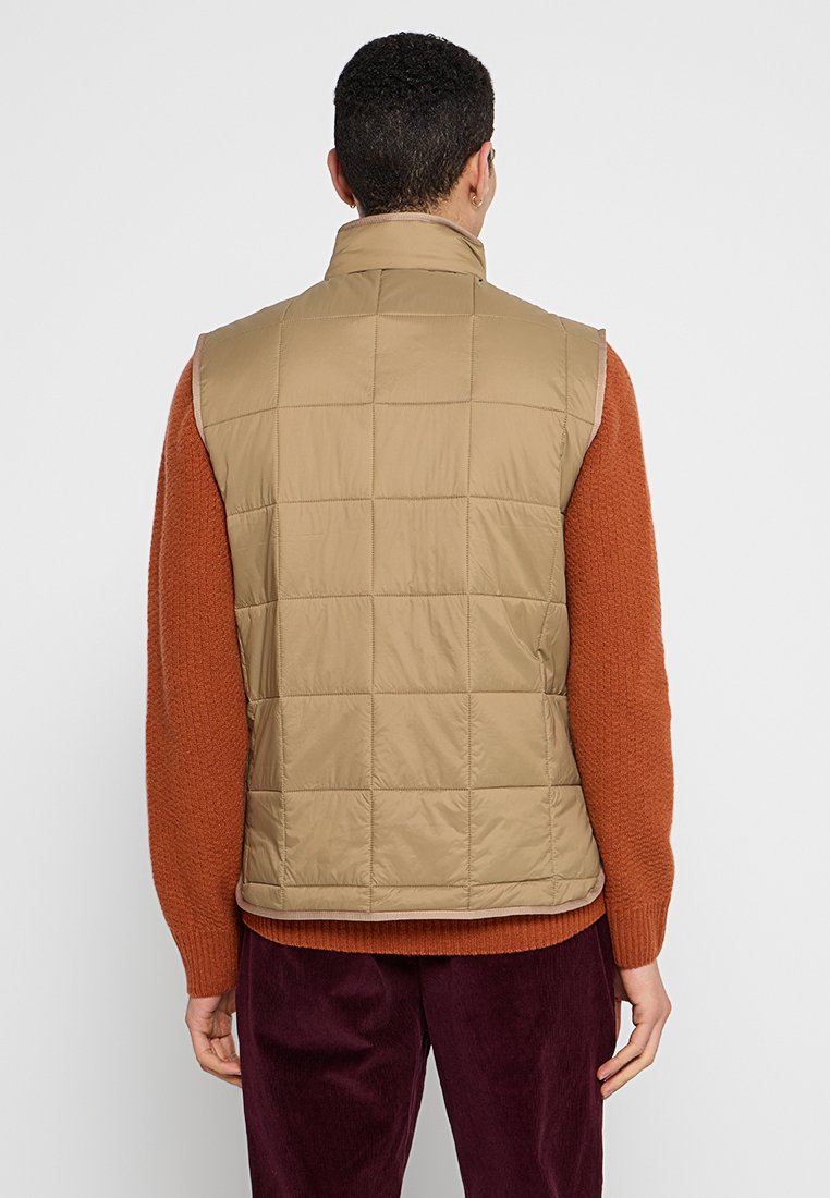 Gilet imbottito trapuntato marrone chiaro con colletto alto, caratterizzato da una trama liscia e dettagli di cucitura minimi. Indossato sopra un maglione arancione.